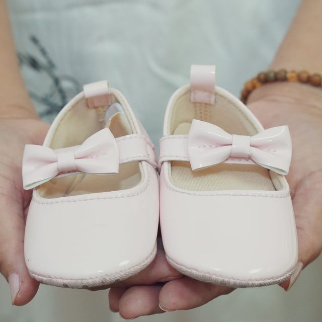 A pair of tiny white baby shoes with bows held in two hands