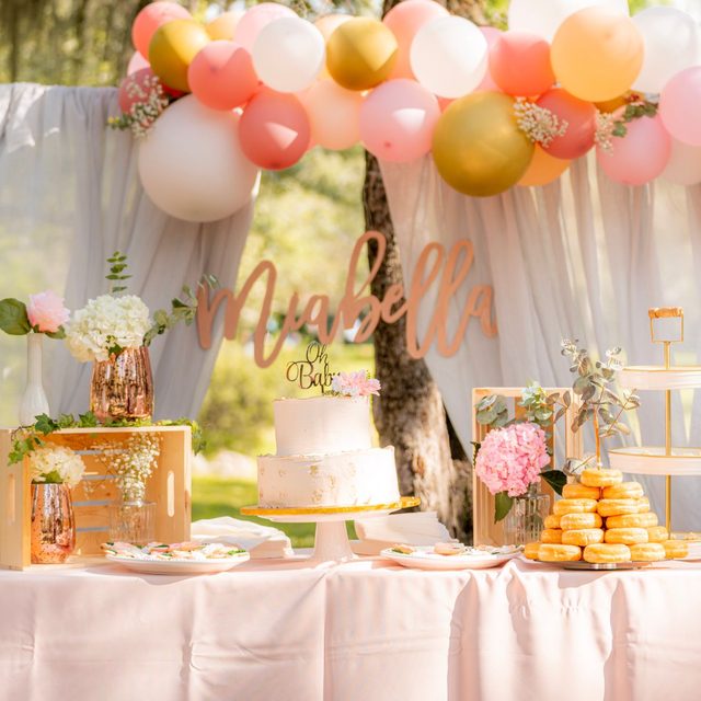 A pastel dessert table set up outdoors under a balloon arch for a baby shower