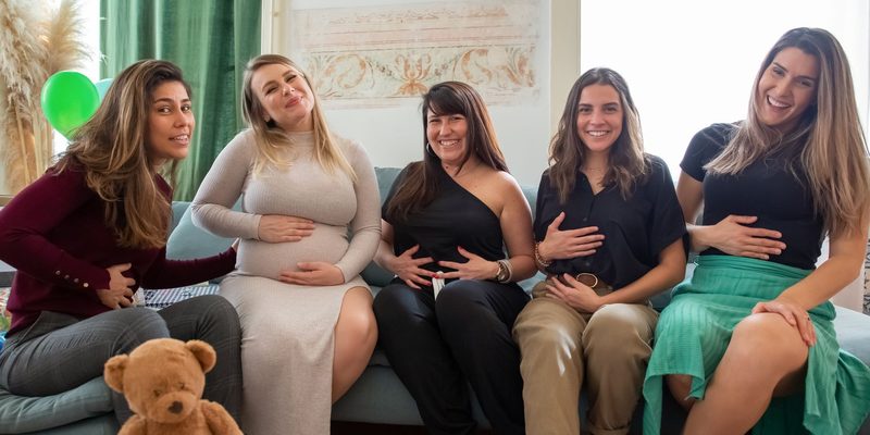 Five women side by side on a sofa at a baby shower, laughing together
