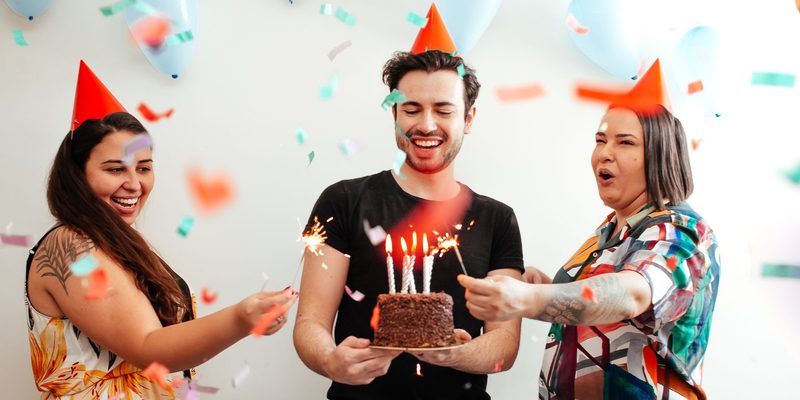 Friends in party hats throwing confetti over someone holding a candlelit cake