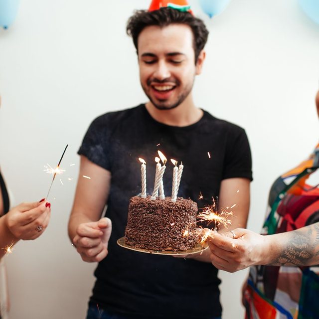 A man in a party hat holding out a chocolate birthday cake lit with sparklers