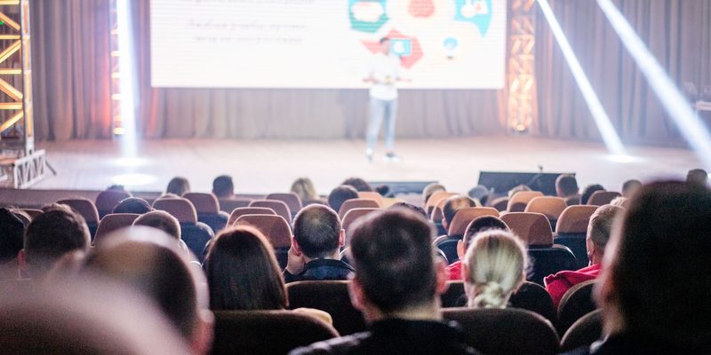 A full auditorium watching a presentation, lit mostly by the screen on stage