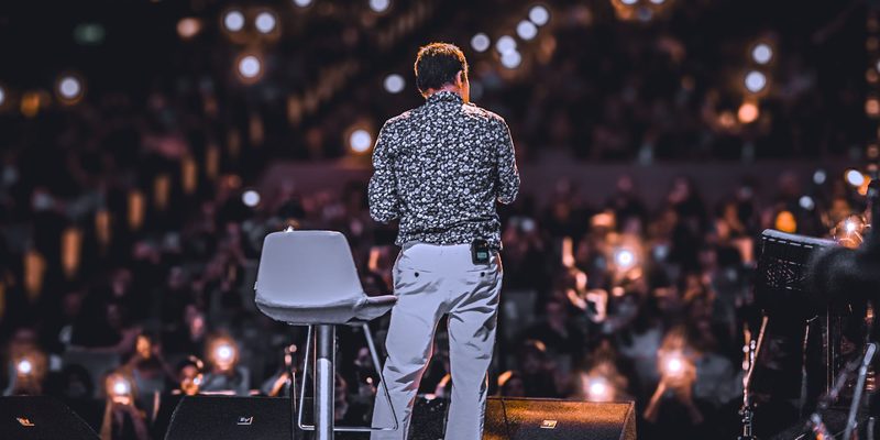 A speaker alone on stage in front of an audience holding up lit phones