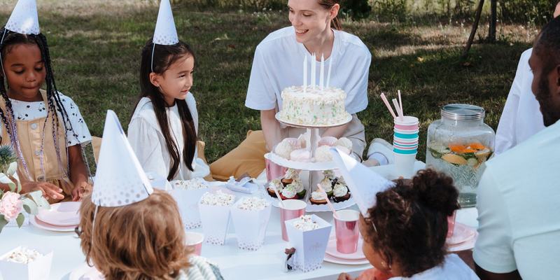 Children in paper party hats crowded around a birthday cake at a garden table