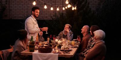 A long garden table of friends eating dinner under strings of bulbs