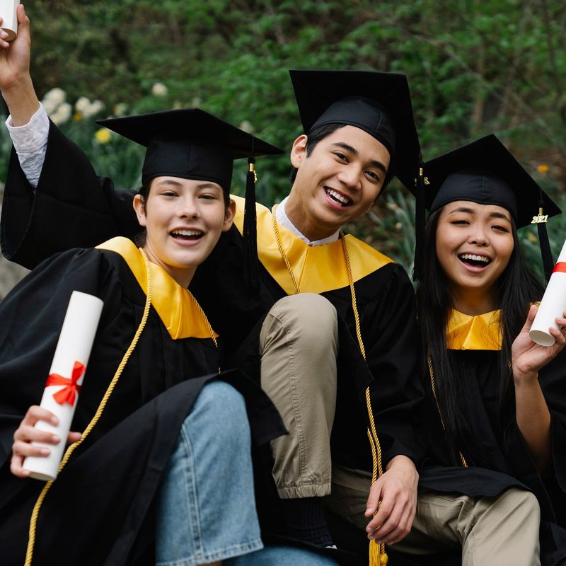 Three graduates in caps and gowns crouching together holding their diplomas