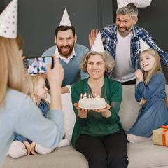 A grandmother holding a birthday cake while her family photographs her on a phone
