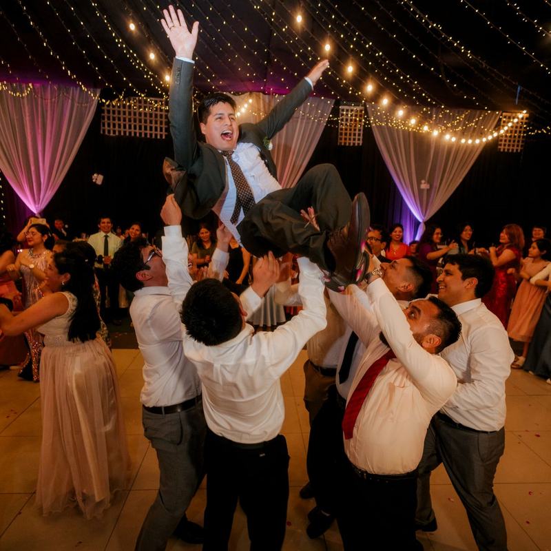 Wedding guests hoisting the groom into the air on a dance floor hung with fairy lights